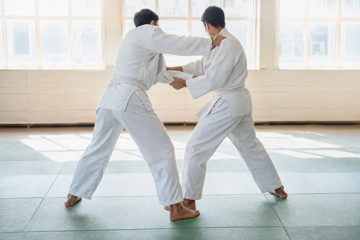 Deux judokas adultes en judogi blanc vus de dos en position de saisie sur un tatami, dojo lumineux à grandes fenêtres