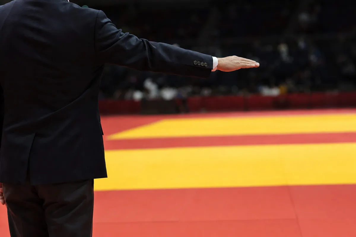 Arbitre de judo en costume sombre annonçant osaekomi bras tendu au bord du tatami de compétition