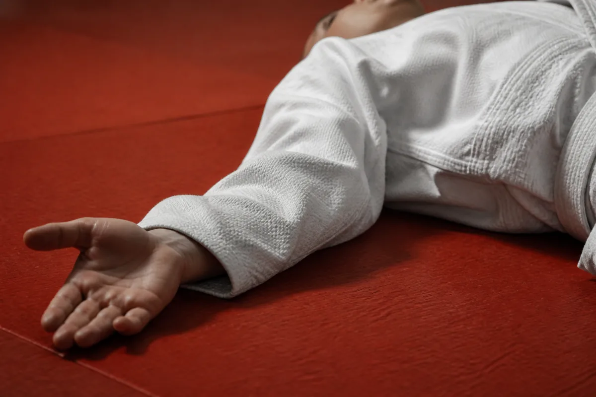 Judoka allongé dos au tatami rouge, épaules à plat et bras tendu en frappe de chute, tissu du judogi froissé sous une lumière rasante
