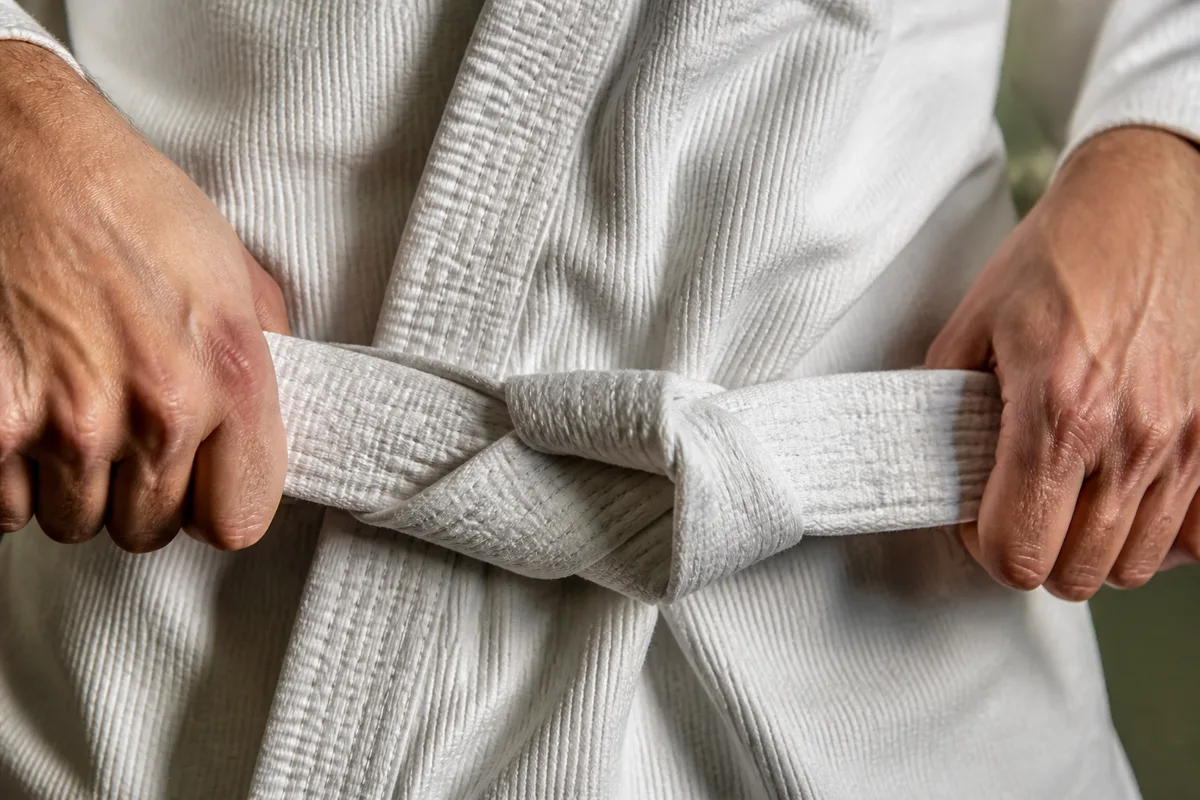 Mains d’un judoka serrant le nœud plat de sa ceinture blanche sur un judogi en coton grainé