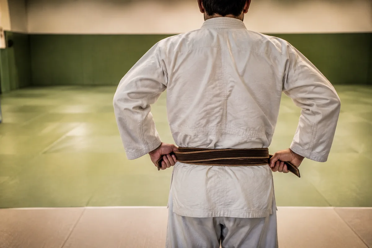 Judoka de dos resserrant sa ceinture marron au bord d’un tatami vert, mains à hauteur de taille